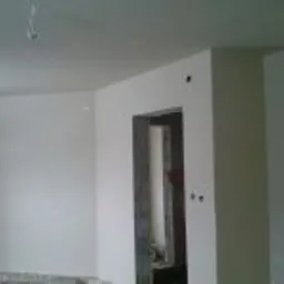 Wnętrze pomieszczenia po nałożeniu gładzi gipsowej, widoczne świeżo wykończone ściany w jasnym odcieniu, sufit z wystającymi przewodami elektrycznymi, otwór drzwiowy z widocznymi śladami po pracach...